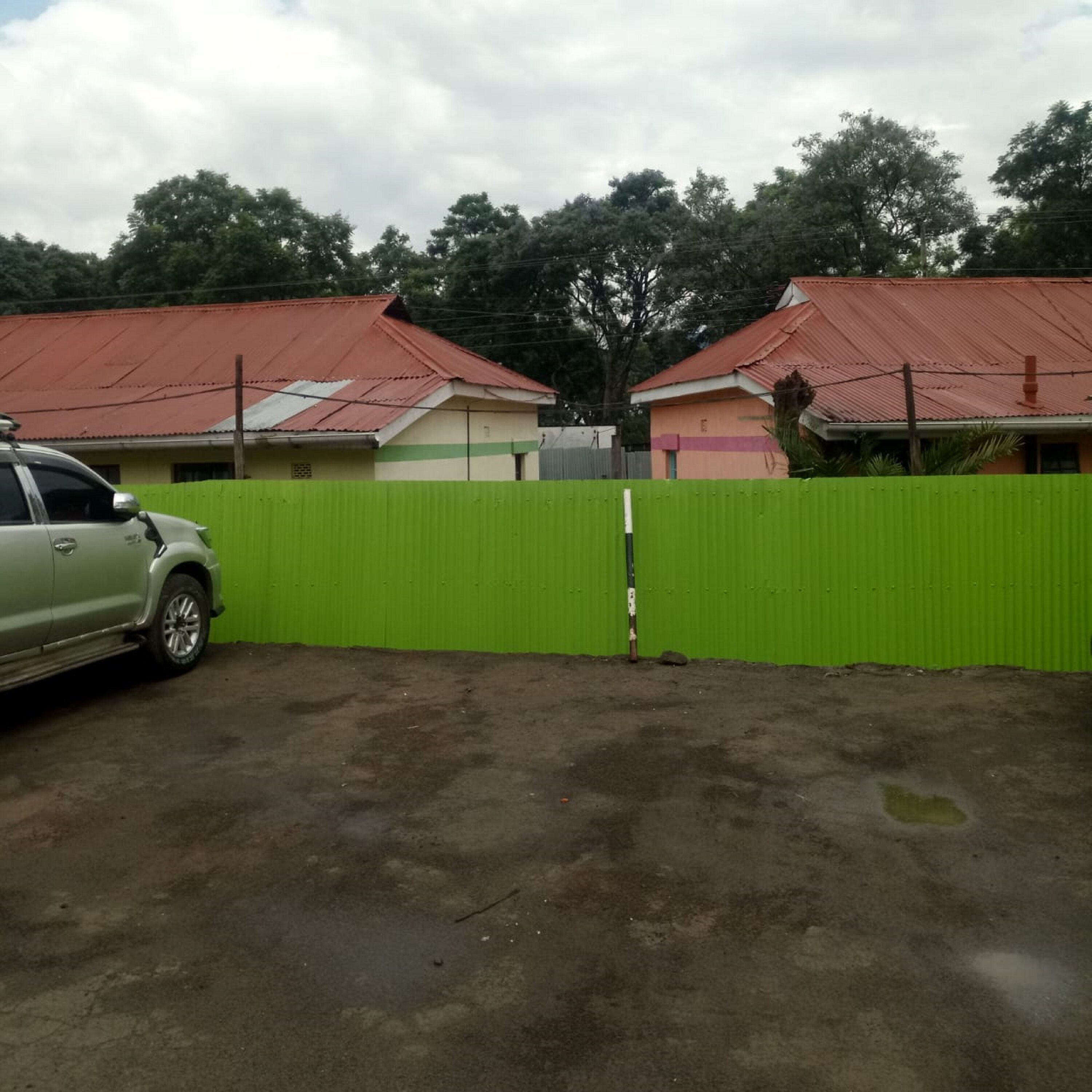 Un área vallada con cerca verde, un coche aparcado y edificios con tejados rojos al fondo.