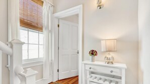 A hallway with a white door, a white console table with a lamp, and a window with blinds. - Bright & Secluded Apartment with Pool Access (Charleston)