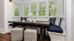 A room with a large window, a wooden table, and two woven ottomans.