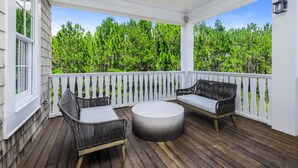 A covered porch with two wicker chairs and a round table, white railings, and a view of green trees.