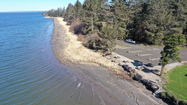 An aerial view of a coastal road with parked vehicles, a parking lot, and dense forest. - Exclusive Bayside RV Site #4 (Aberdeen)