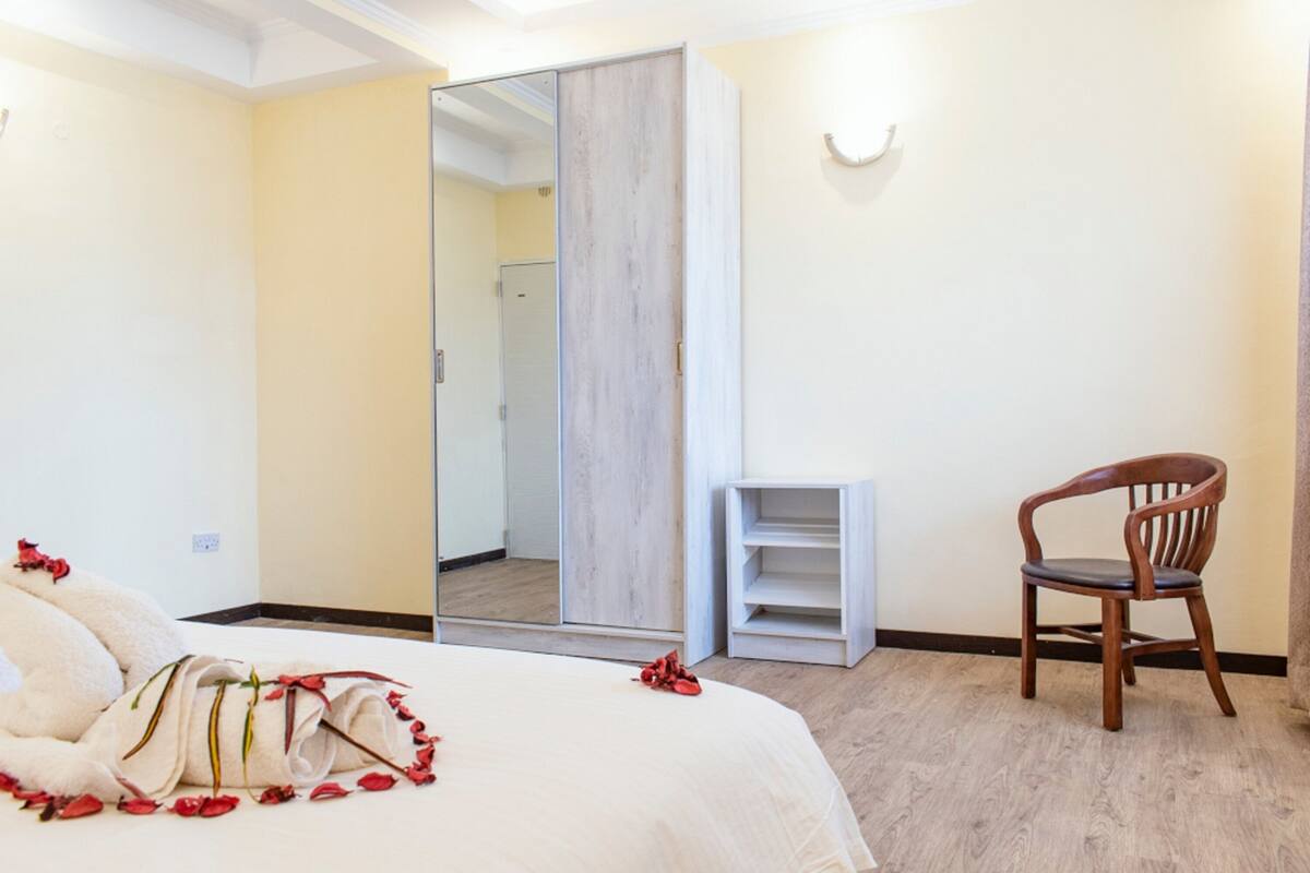 A hotel room with a bed, a wooden chair, a mirror, and a small white shelf.