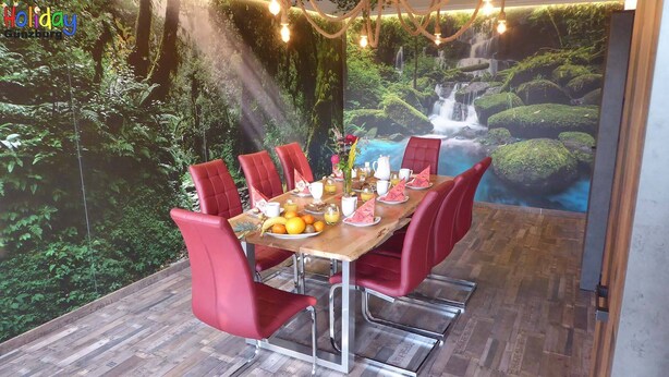 A dining area with a mural of a waterfall and forest, red chairs, and a wooden table set for breakfast. - Apartment Jungle Adventure (Günzburg)