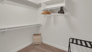 Habitación de esquina con paredes blancas, alfombra color beige, un cesto de basura pequeño, una silla de metal negro y un perchero con ropa.