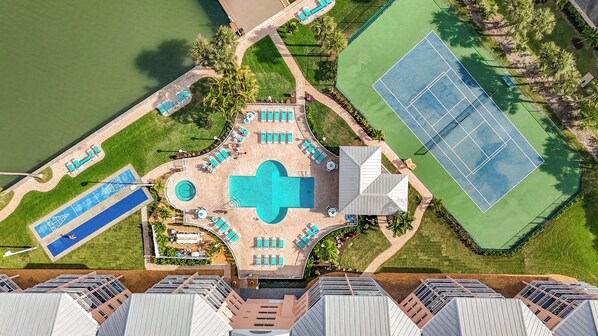Vue aérienne d’un complexe hôtelier doté d’une piscine centrale, de nombreuses chaises longues, d’un court de tennis et d’un terrain de jeu de palet.