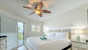 A bedroom with a bed, a nightstand, a lamp, a ceiling fan, and a view of the outdoors. - Beach Luxury Resort | Bay Views (St. Pete Beach)