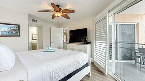 A bedroom with a bed, a ceiling fan, a TV, and a sliding glass door to a balcony. - Beach Luxury Resort | Bay Views (St. Pete Beach)