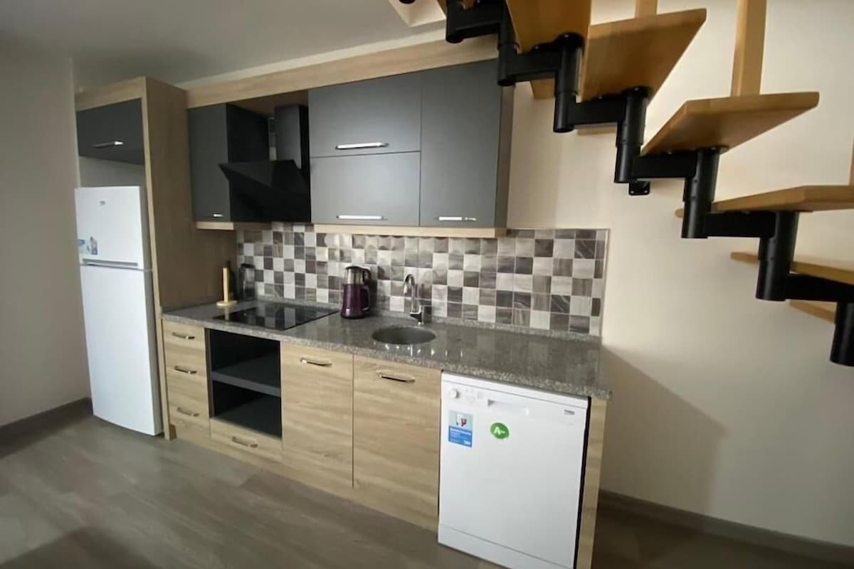 A modern kitchen with grey cabinets, a checkered backsplash, a built-in dishwasher, and a unique wooden staircase.