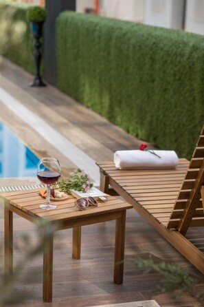 Una poltrona da relax in legno con un asciugamano, un bicchiere di vino rosso e occhiali da sole su un tavolino vicino a una piscina.