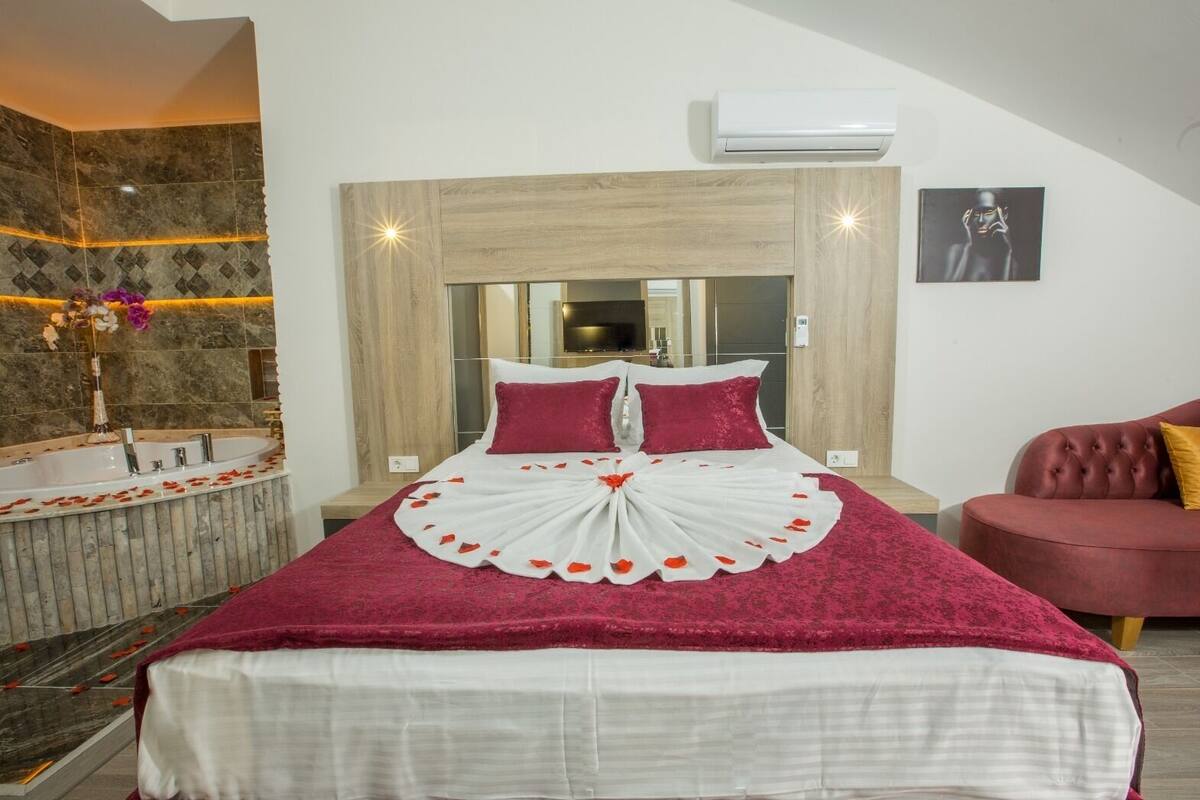 A bedroom with a large bed, a wooden headboard, a red and white floral bedspread, a red armchair, and a bathtub with rose petals.