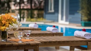 A wooden table with a vase of flowers, a glass of water, and a martini glass. There are towels and a red rose on a bench by a pool.