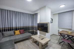 A modern living room with a grey sofa, a wooden coffee table, a mounted TV, and a dining area with a table and chairs. - Elanis Suites (Antalya)