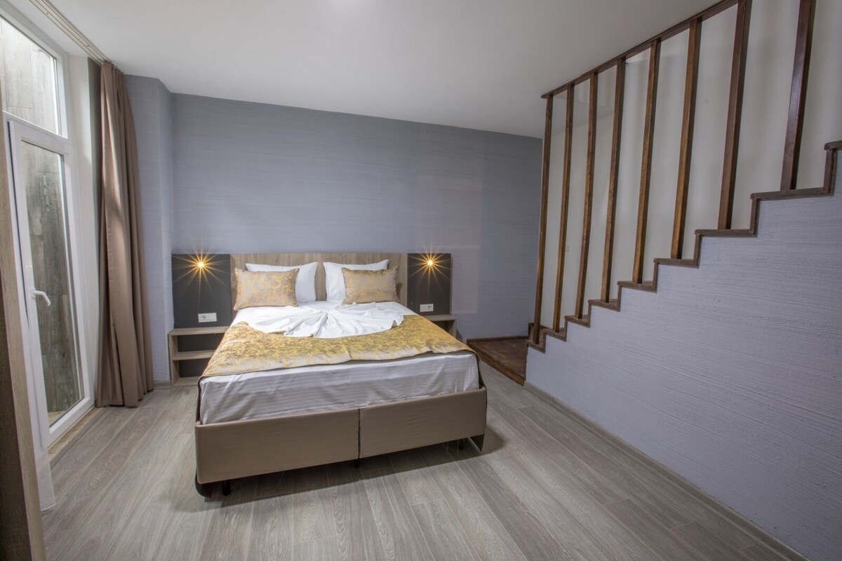A bedroom with a bed, nightstands, a staircase, and a window with curtains.