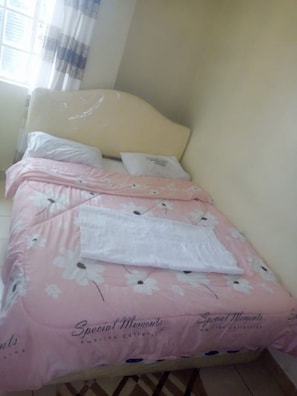 A neatly made bed with a floral bedspread and pillows, a headboard, and a window with curtains. - Senior Chief Wambugu Palace (Marua)