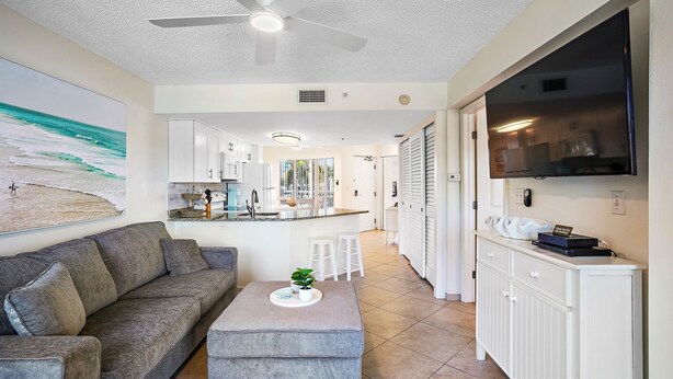 Ein modernes Wohnzimmer mit grauem Sofa, grauen Hocker, Deckenventilator, Flachbildfernseher und einer Küchenzeile mit Bar.