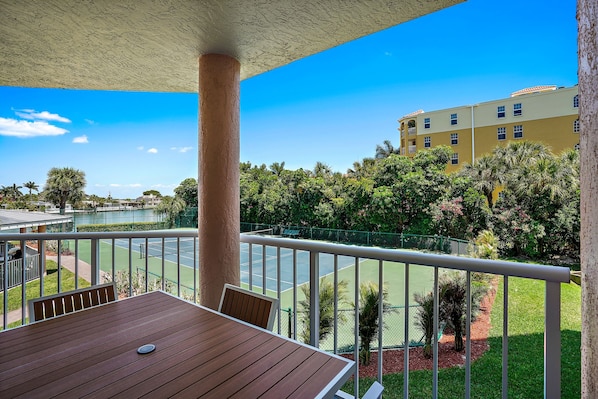 A balcony with a wooden table and chairs, overlooking a tennis court and lush greenery.