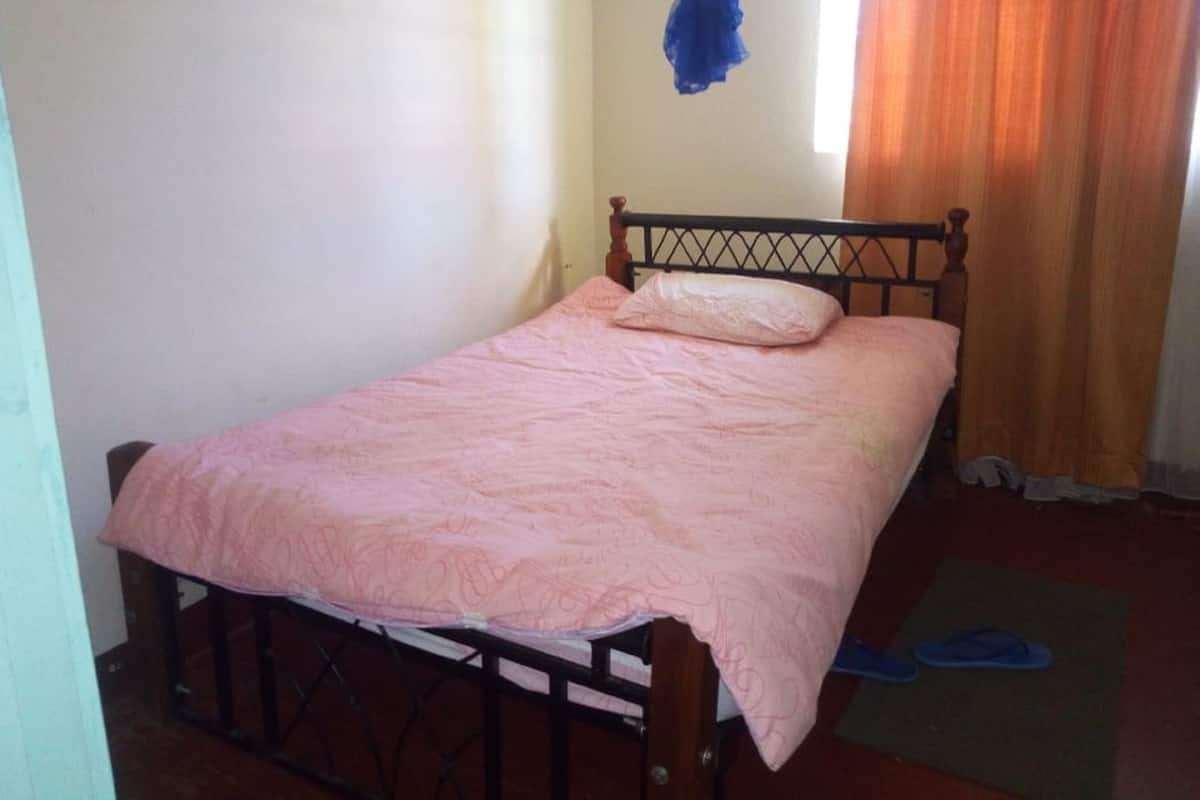 Una cama individual con estructura de madera y colcha rosa en una habitación con pared blanca lisa y ventana con cortinas.
