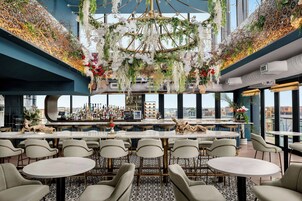 A modern bar area with a high ceiling, decorative hanging plants, and a view of the cityscape through large windows.