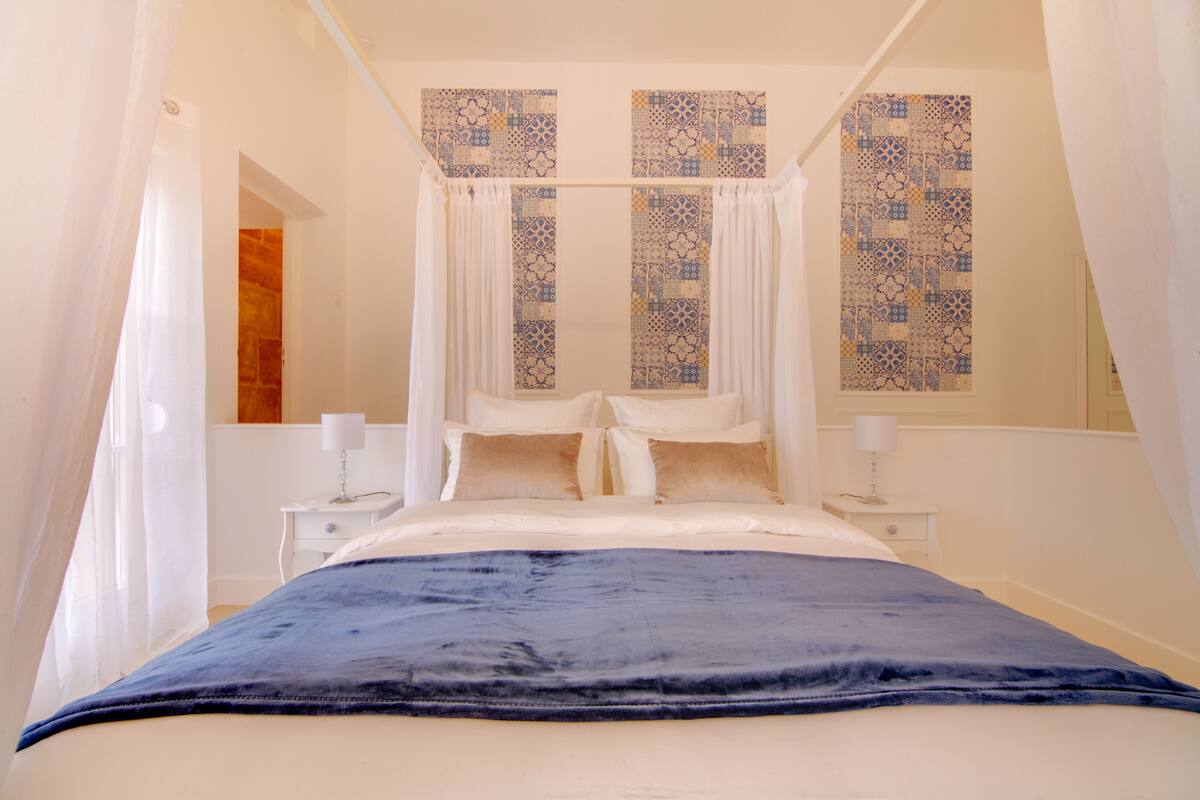 A bedroom with a four-poster bed, white curtains, and patterned wall decorations.