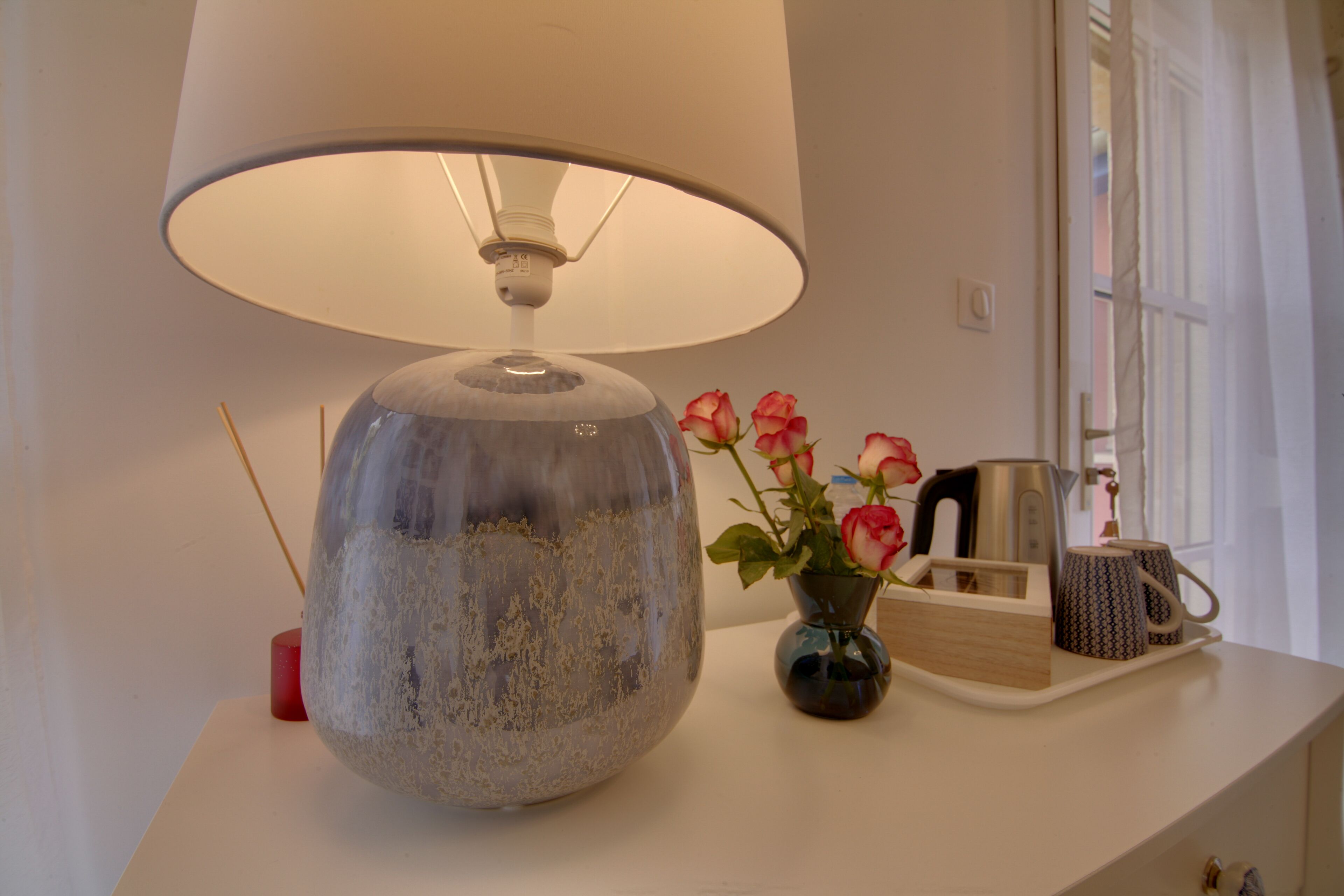 Une lampe avec un grand abat-jour, un vase rempli de roses et un plateau avec une théière et des tasses, le tout posé sur une table blanche près d’une fenêtre.