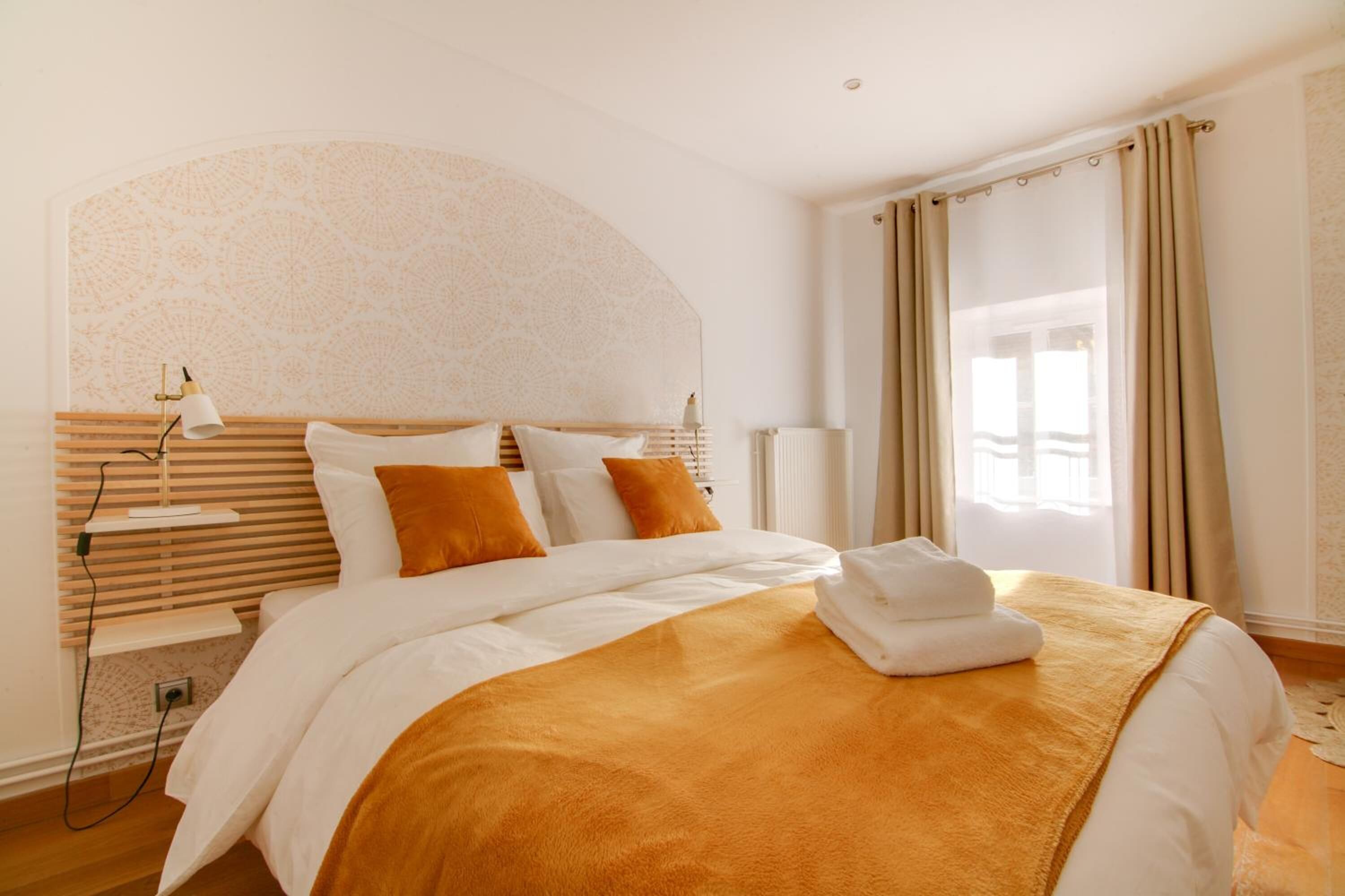 Una habitación de hotel con cama, cabecero, mesita de noche con lámpara y ventana con cortinas.