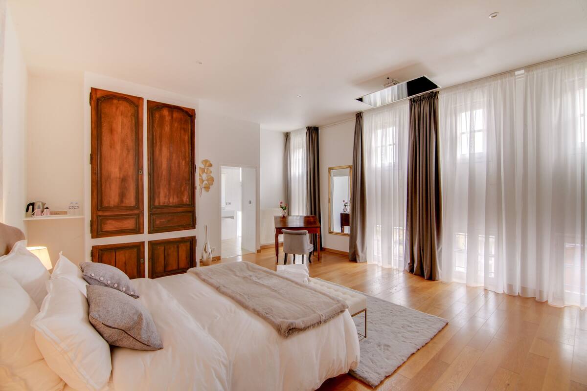 A spacious bedroom with a large bed, a wooden wardrobe, a desk, and a large window with curtains.