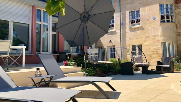 Un patio al aire libre con sillones de descanso y un paraguas grande, rodeado por un edificio de piedra con varias ventanas y una puerta.