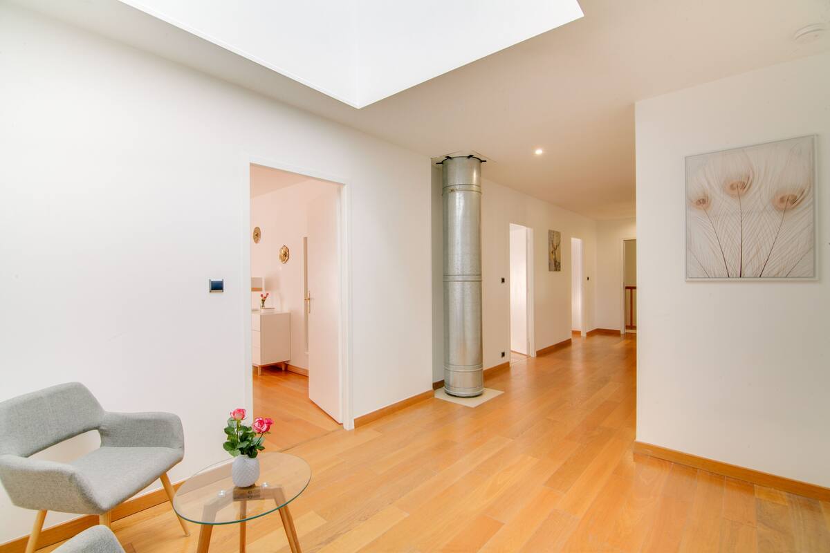 A spacious hallway with wooden flooring, a modern chair, a glass table with flowers, and a tall metallic column.