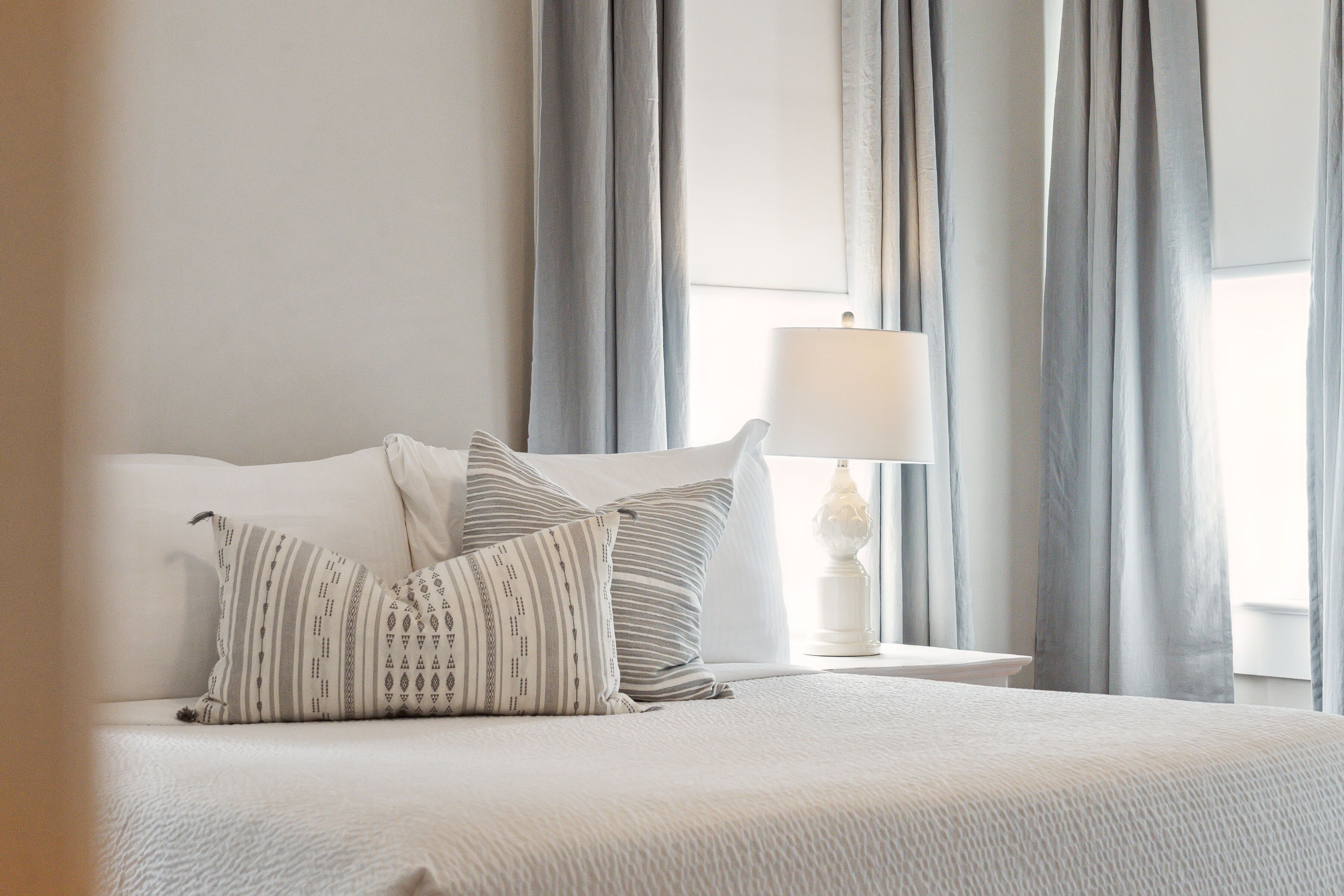 A neatly made bed with a white comforter and two pillows, a bedside table with a lamp, and light gray curtains.