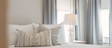 A neatly made bed with a white comforter and two pillows, a bedside table with a lamp, and light gray curtains.