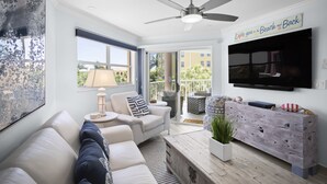 Uma sala de estar espaçosa com um sofá branco, uma mesa de centro e um ventilador de teto. Uma TV está fixada na parede, e há uma varanda com mobiliário de exterior.