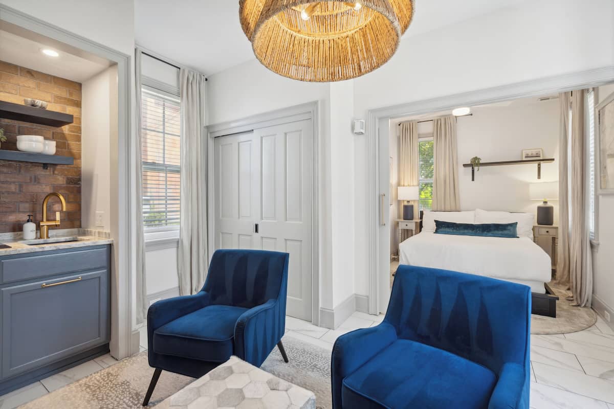 A modern bedroom with a large bed, two blue armchairs, a white dresser, and a kitchen area with a brick backsplash.