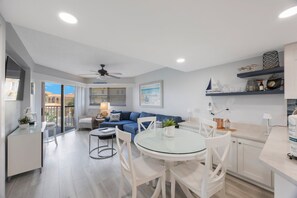A modern living room with a blue sofa, a glass dining table, white chairs, and a ceiling fan.