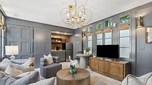 Una sala moderna con un sofá gris, una mesa redonda con una planta, un televisor y una cocina al fondo.