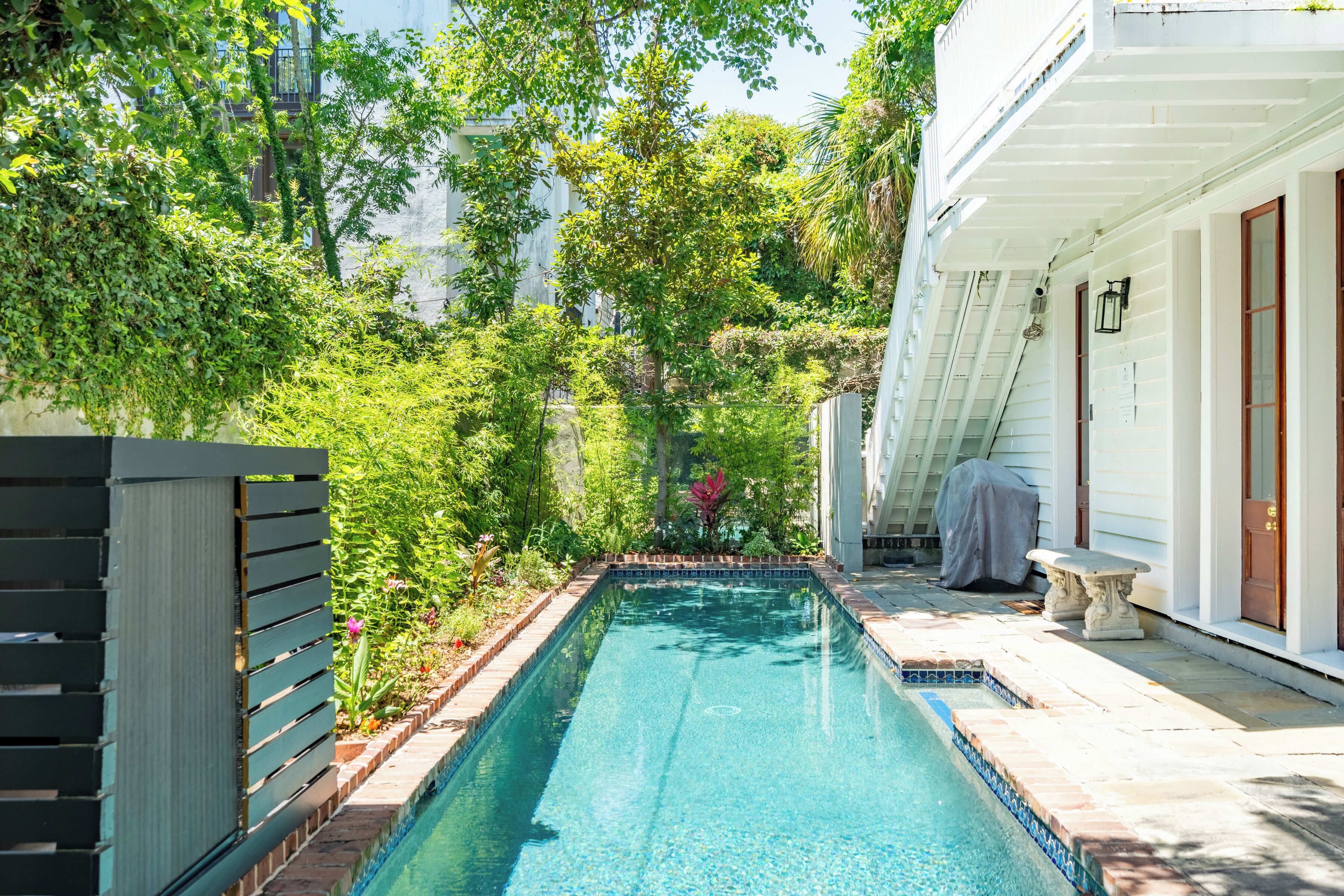 Une piscine rectangulaire bordée de carreaux, entourée d’une végétation luxuriante et d’un bâtiment blanc doté d’un banc abrité et d’une porte.