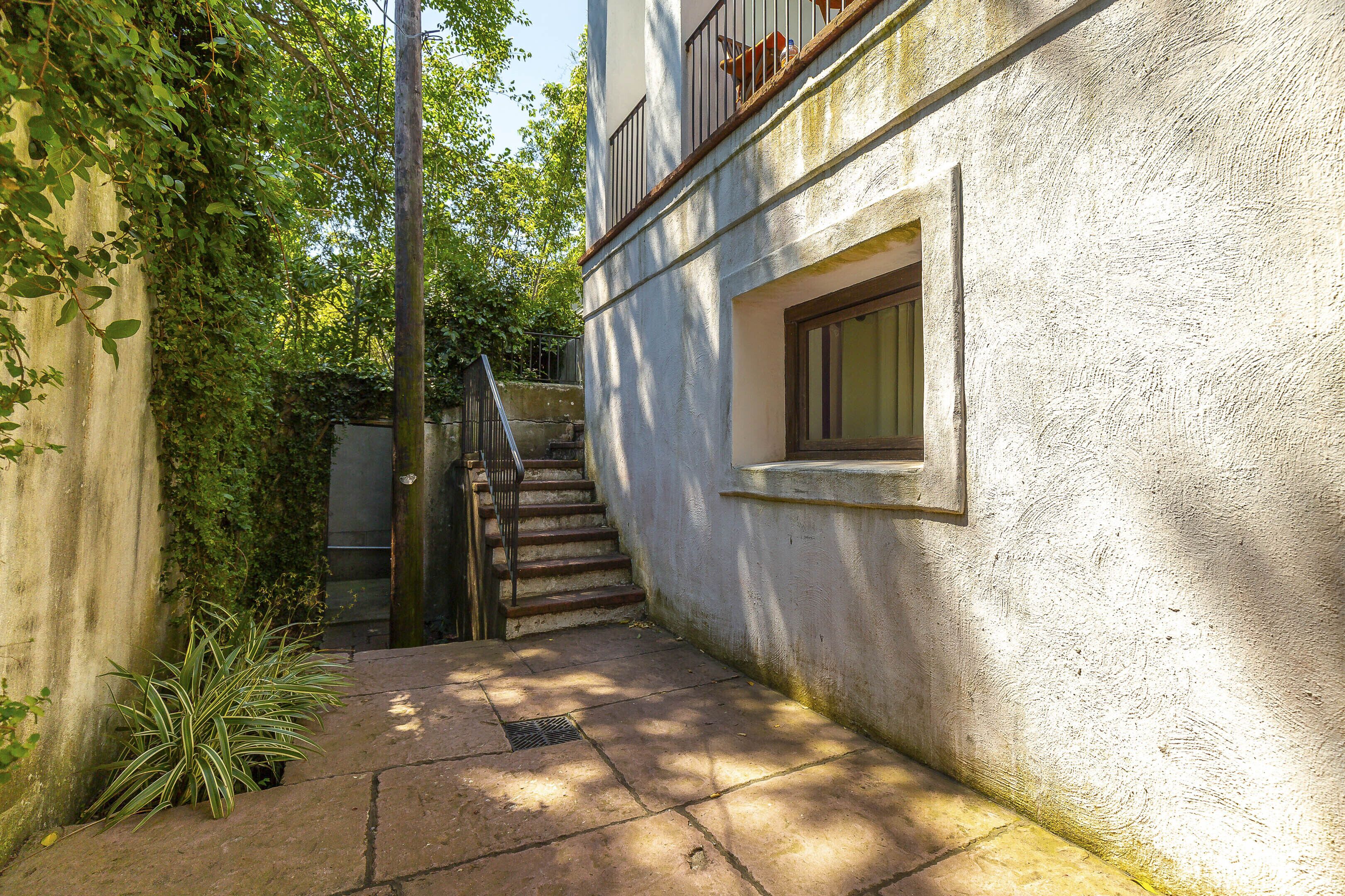 A narrow outdoor corridor with a concrete floor, a small window, and a staircase leading to a balcony.