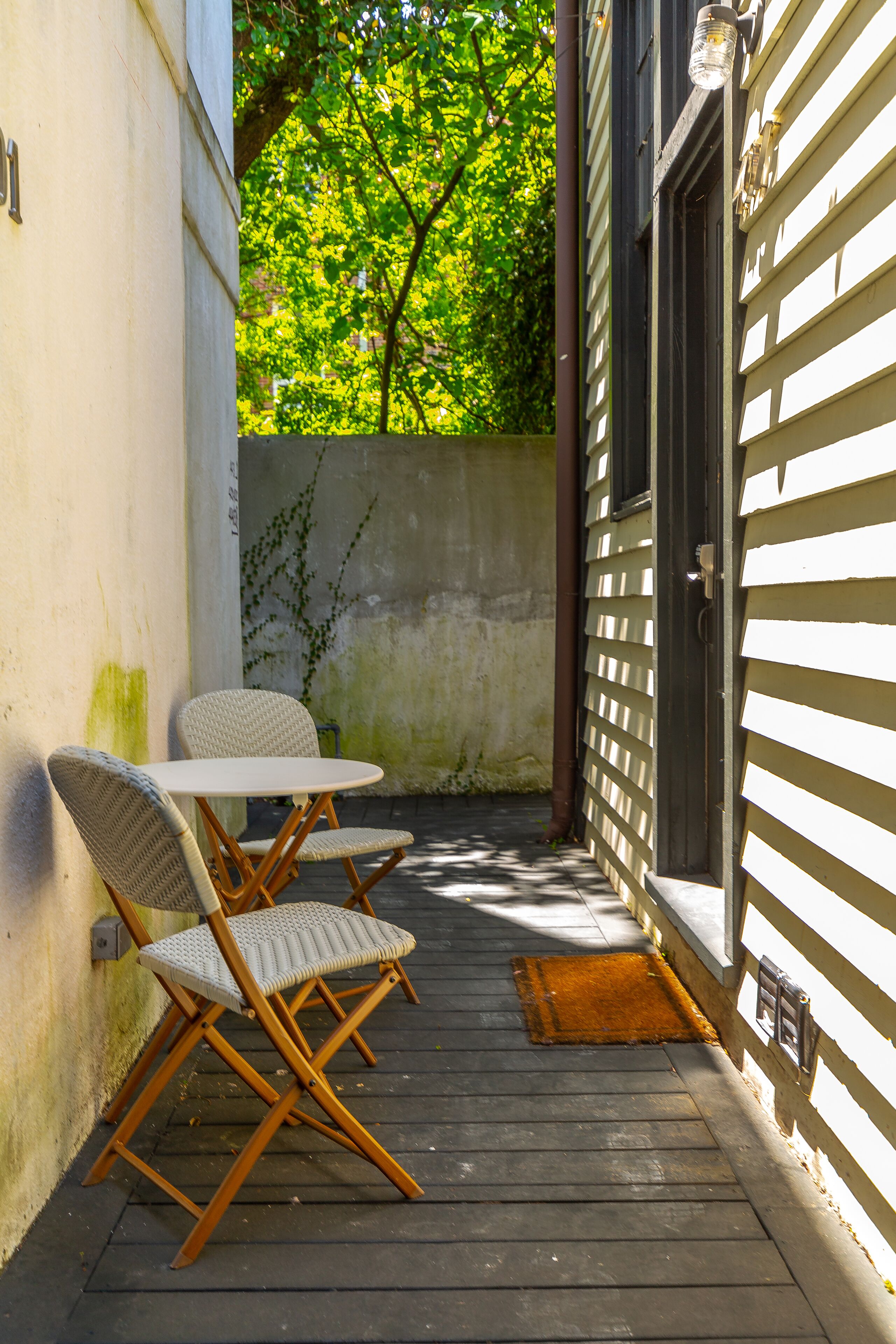 A small patio with a table and two chairs, a wooden door, and a concrete wall with some greenery.