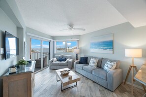 Un salon spacieux avec un canapé gris, une table basse et un téléviseur. Il y a un balcon avec vue sur la mer.
