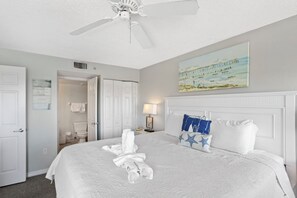 Un lit bien fait, avec du linge de lit blanc, un ventilateur de plafond, un tableau représentant une jetée, et une salle de bain visible à travers une porte ouverte.