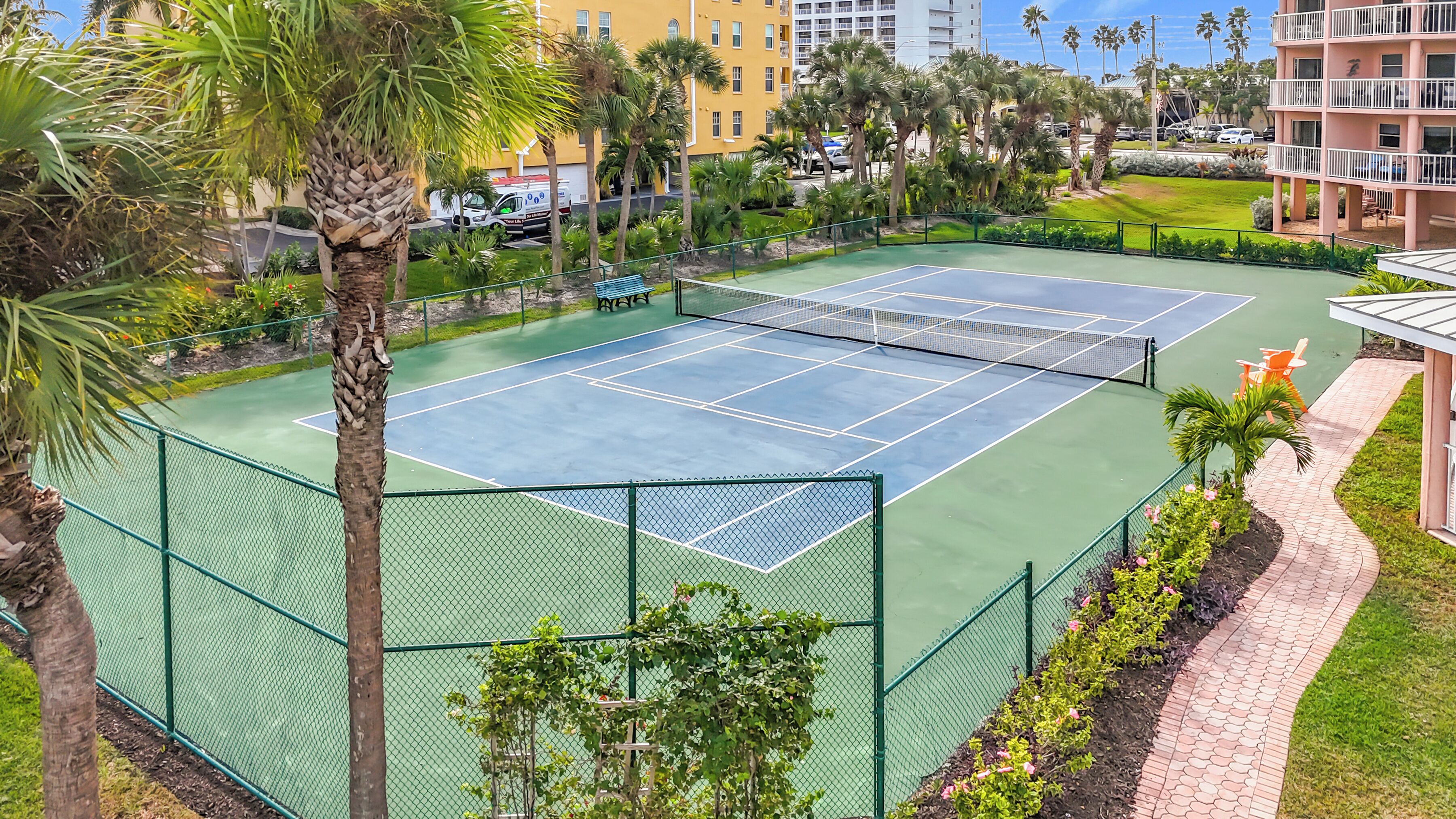 A tennis court surrounded by a green fence, palm trees, and a pathway leading to a building with a balcony.