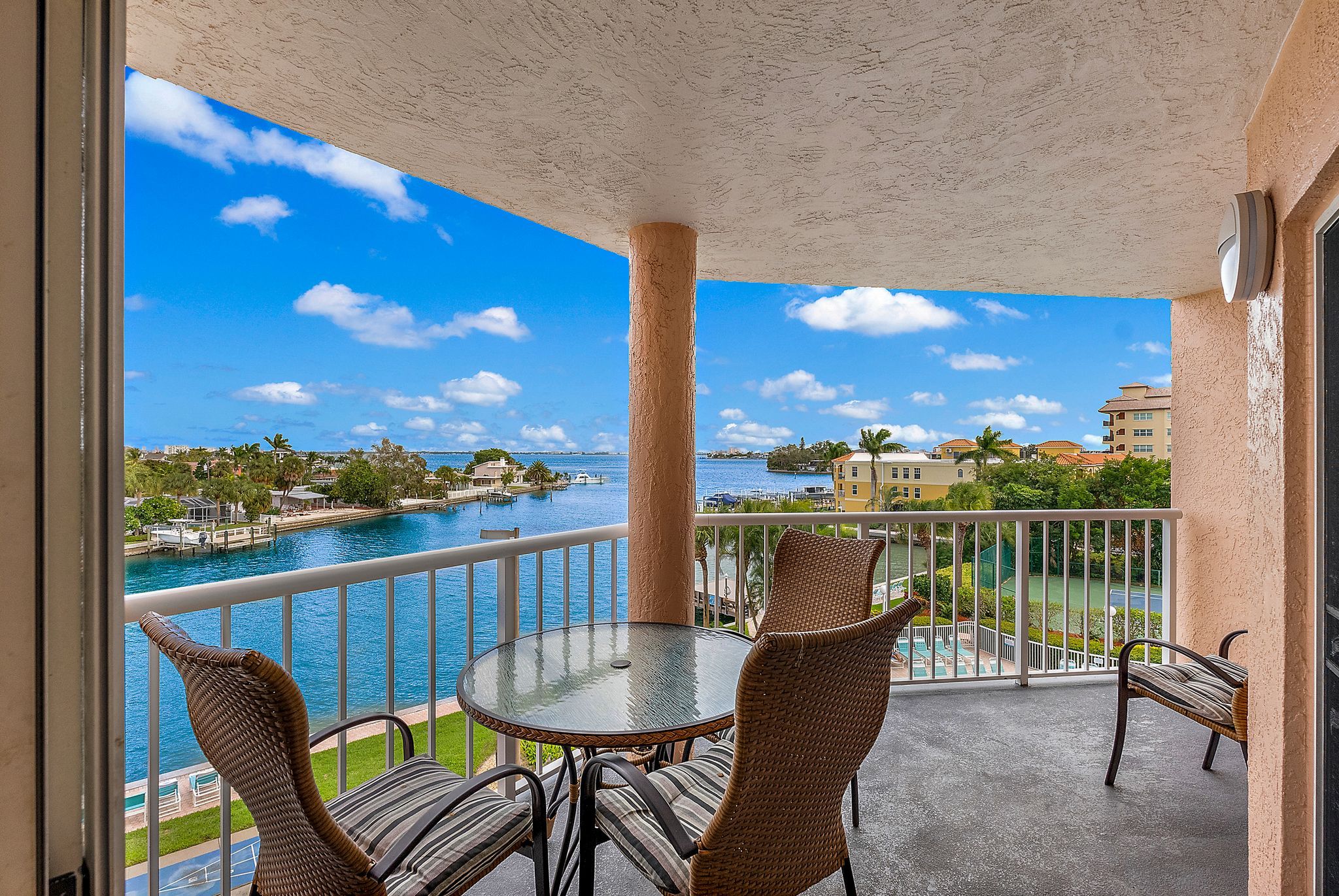 A balcony with a glass table and wicker chairs, overlooking a body of water and a marina.