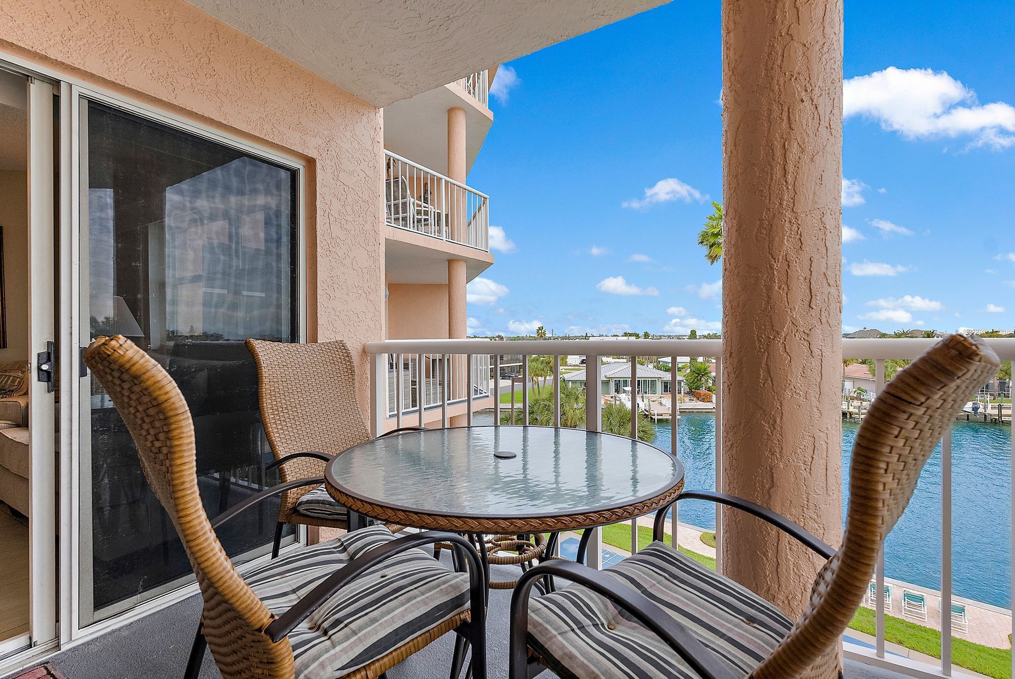 A balcony with wicker chairs and a glass table, overlooking a body of water and a resort area.