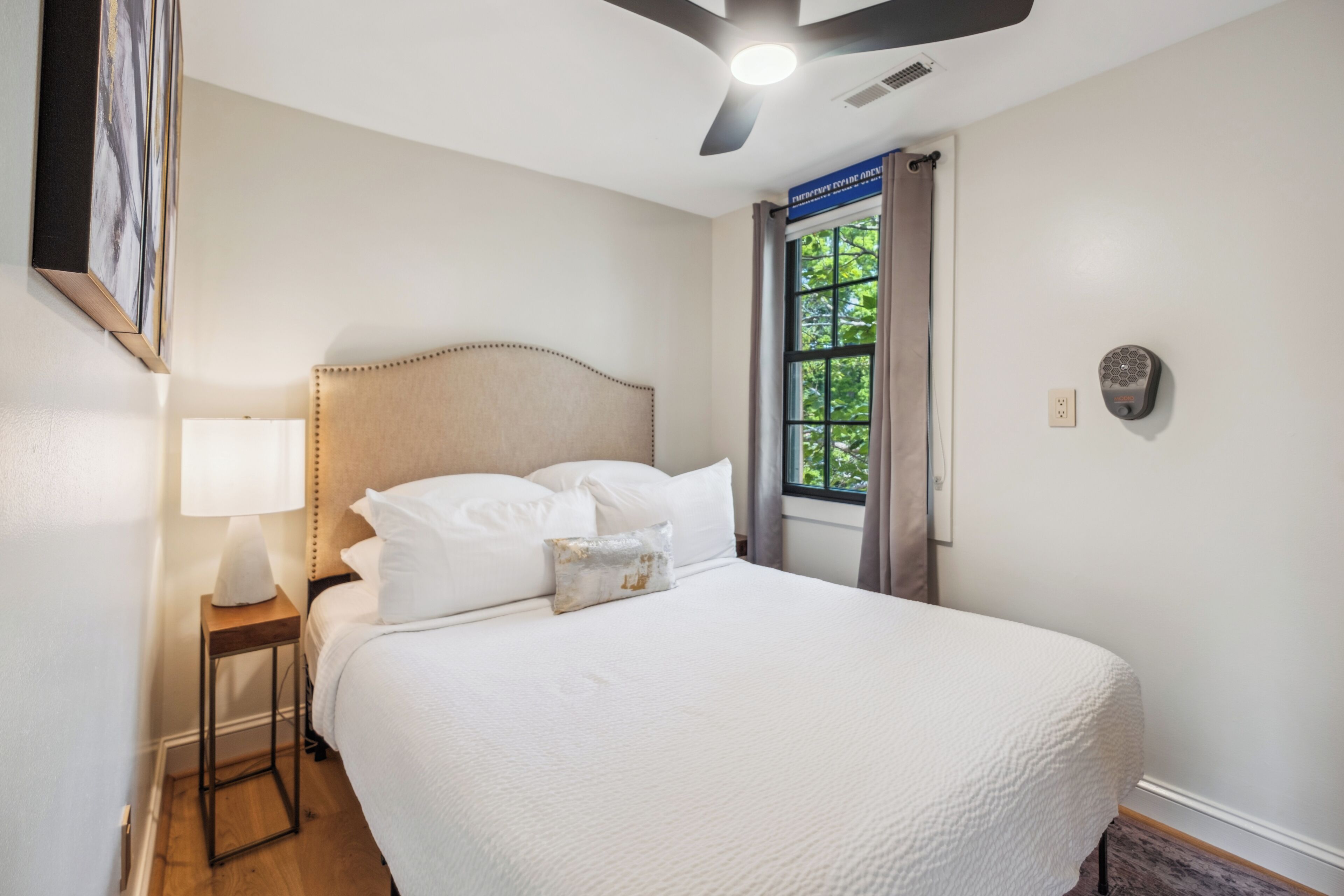 Una cama bien hecha con cabecera, una lámpara de noche y un ventanal con cortinas.