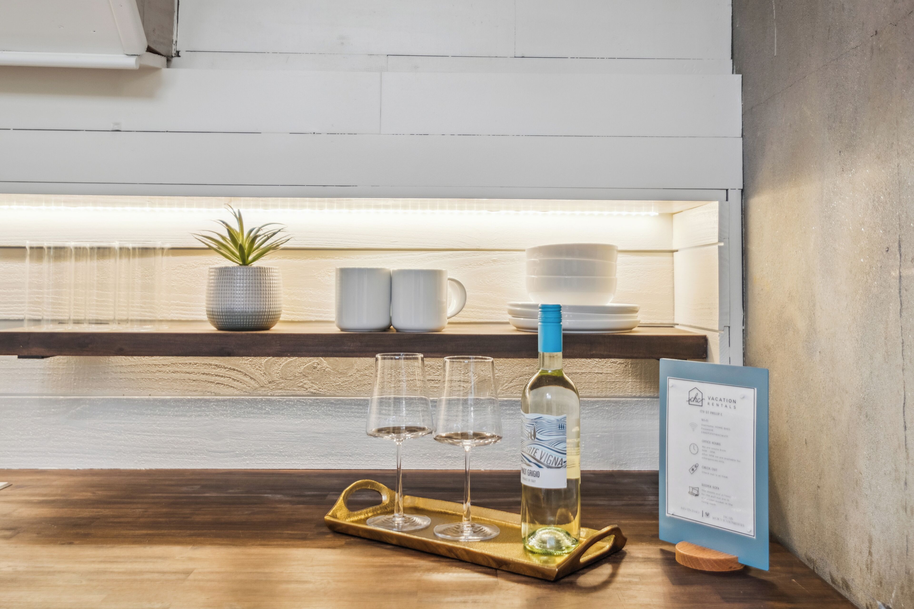 A wooden table with a tray holding a bottle of wine and three glasses, a potted plant, and a menu board.