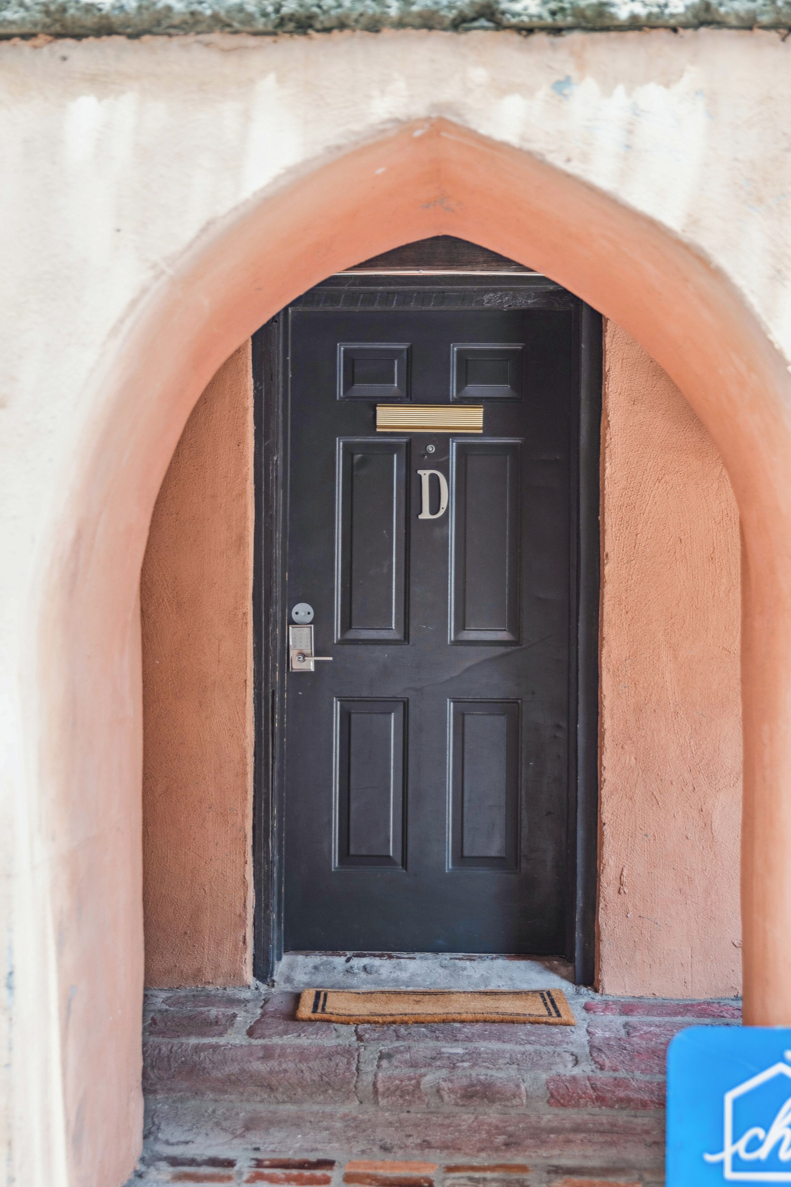 Uma porta preta com o símbolo 'D', emoldurada por uma porta em arco, diante de uma parede texturizada.