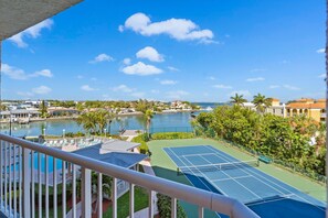 A tennis court with a pool in the background, surrounded by greenery and buildings. - Tranquil Bayside Condo | Steps to Beach (St. Pete Beach)