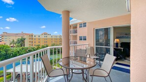 A balcony with a glass table and chairs, overlooking a swimming pool and a hotel building. - Coastal Escape | Pool, Tennis, + Shuffleboard (St. Pete Beach)