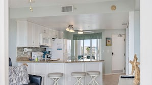 A modern kitchen with white cabinets, a central island with stools, a ceiling fan, and a view to the outside. - Coastal Escape | Pool, Tennis & Pickleball (St. Pete Beach)