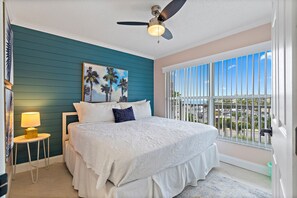 A bedroom with a bed, a ceiling fan, a nightstand with a lamp, a painting of palm trees, and a view of the city through large windows.