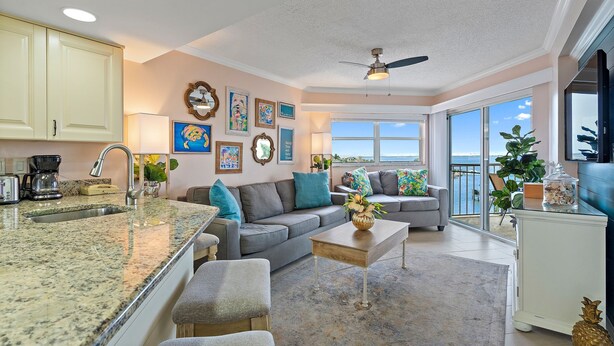 A modern kitchen with granite countertops, a built-in oven, and a large island. There is a gray sofa and a dining table with chairs. The room has large windows with a view of the ocean. - Sun, Sand & Style | Ideal Beach Retreat (St. Pete Beach)